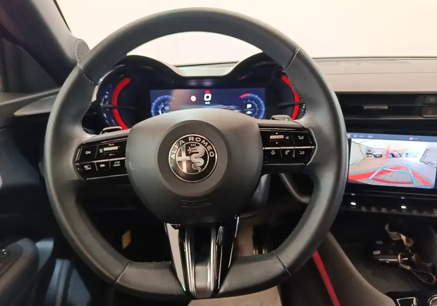 Vue intérieure centrée sur le volant cuir noir d'Alfa Romeo Junior 2024 avec tableau de bord numérique et écran de recul.