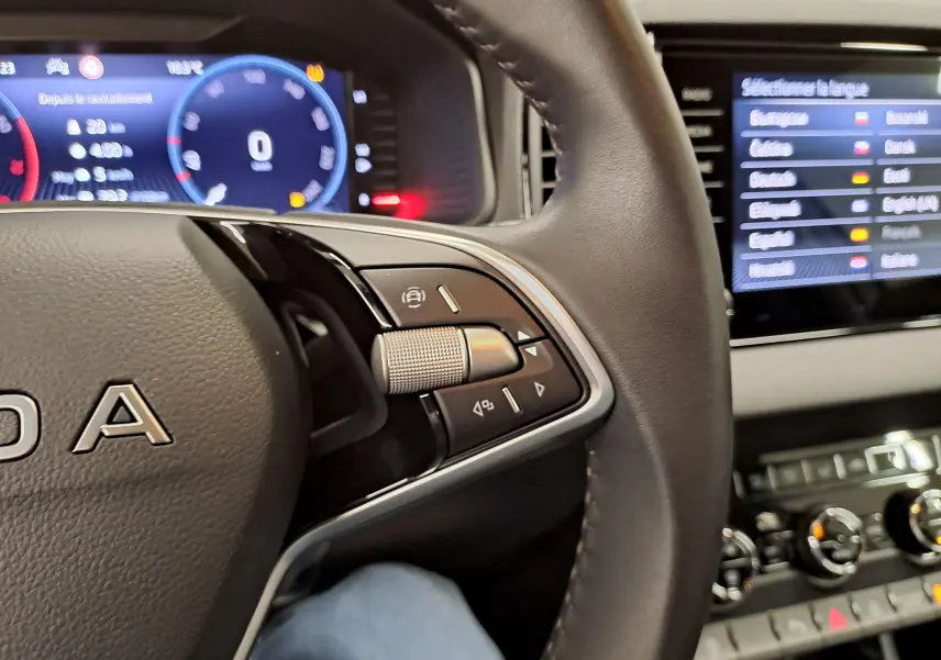 Gros plan sur le volant et le tableau de bord numérique d'une Skoda Karoq blanc lune, version 2025, avec commandes au volant visibles.