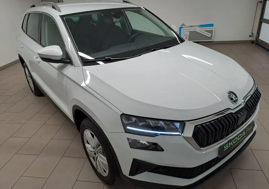 Vue 3/4 avant droit d’un Skoda Karoq blanc lune 2025 avec phares LED allumés et calandre noire distinctive.