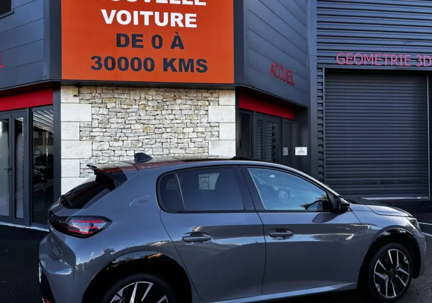 Peugeot 208 Hybrid 136 eDCS6 GT gris foncé vue de profil côté droit devant un bâtiment avec panneau orange.