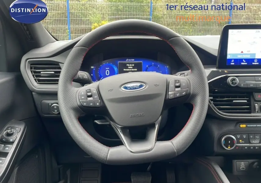 Vue intérieure centrée sur le volant perforé avec coutures rouges du Ford Kuga 2024, tableau de bord digital et écran tactile visibles.