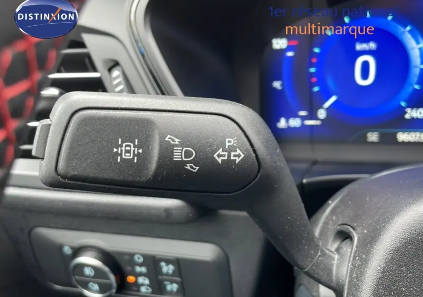 Gros plan sur la manette de clignotant noire à gauche du volant, tableau de bord numérique bleu en arrière-plan.
