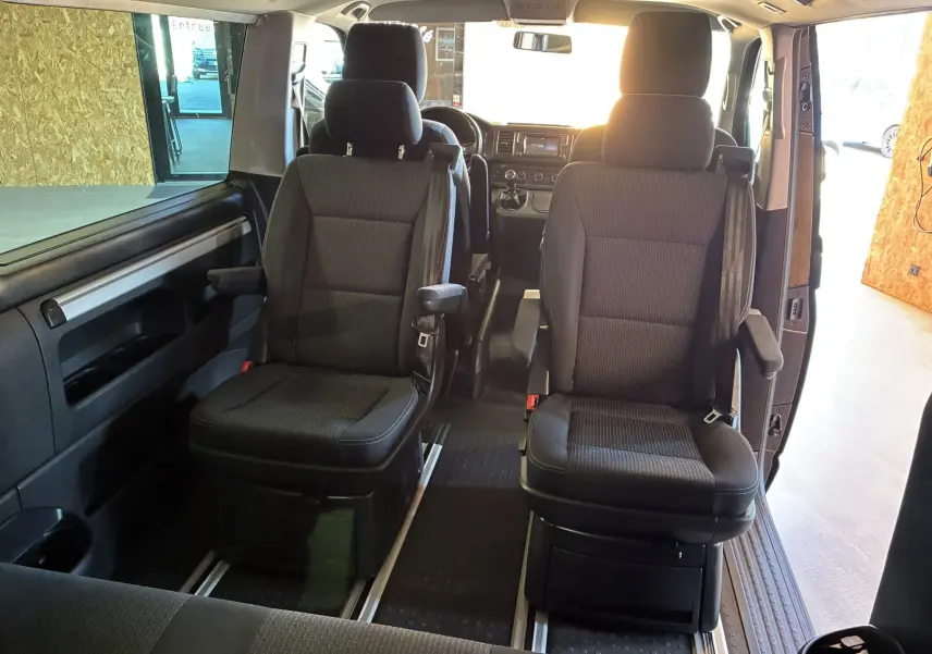 Vue intérieure avant du Volkswagen Multivan T6 noir, montrant les sièges avant individuels avec accoudoirs et tableau de bord.