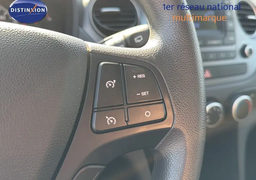 Gros plan sur les commandes de régulateur de vitesse au volant d'une Hyundai i10 blanche, intérieur visible en arrière-plan.