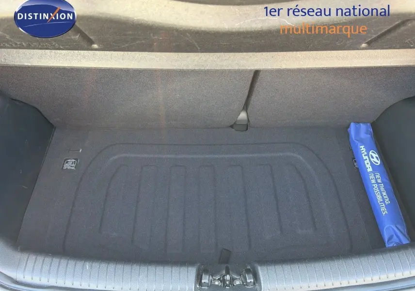 Coffre ouvert d'une Hyundai i10 blanche, vue de dessus, avec tapis de coffre noir et trousse bleue Hyundai sur le côté droit.