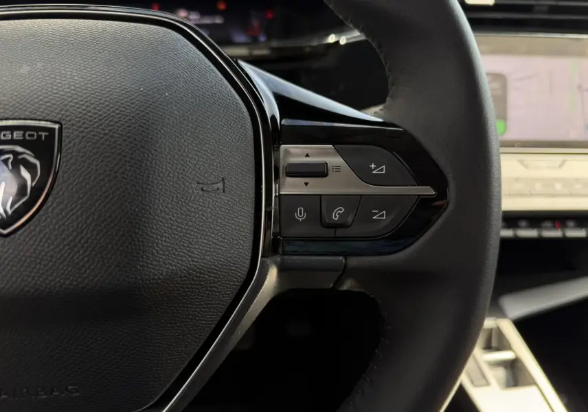 Gros plan sur le volant cuir noir de la Peugeot 308 2025 avec commandes intégrées et tableau de bord numérique en arrière-plan.