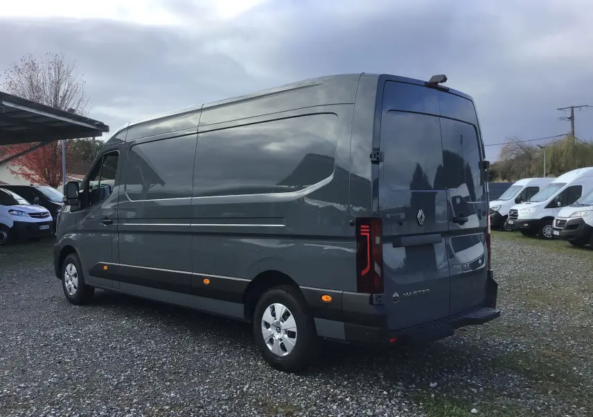Vue 3/4 arrière droite d’un Renault Master gris urbain stationné sur un sol gravillonné avec plusieurs fourgons blancs en arrière-plan.