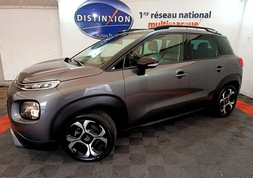 Vue côté gauche 3/4 avant d'un Citroën C3 Aircross gris métallisé avec jantes distinctives et barres de toit noires.