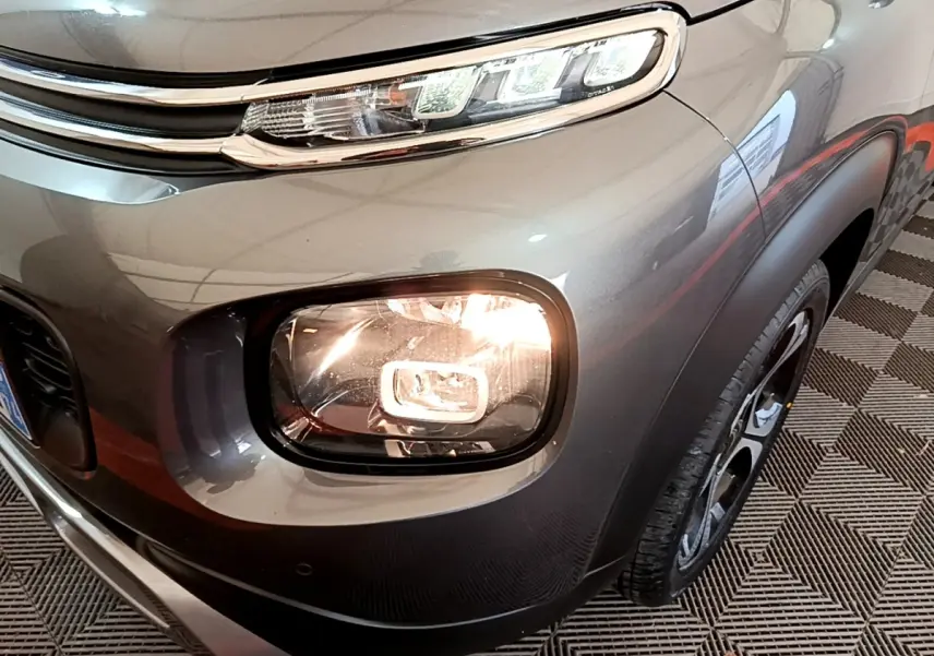 Gros plan sur le phare avant droit allumé d'un Citroën C3 Aircross gris métallisé, avec jante noire visible.