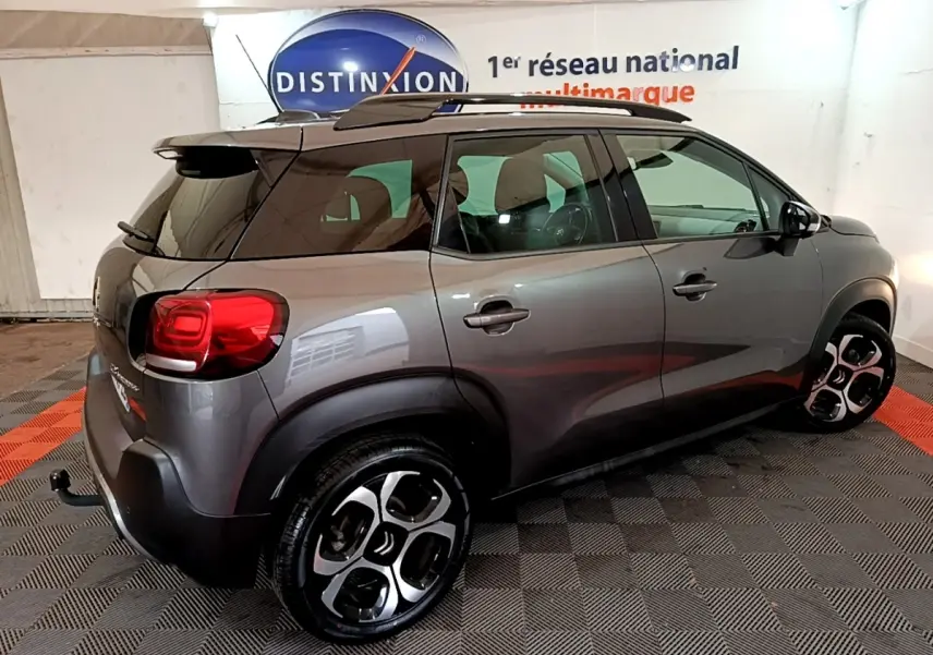 Vue 3/4 arrière droite d'un Citroën C3 Aircross gris métallisé avec barres de toit noires en intérieur showroom.