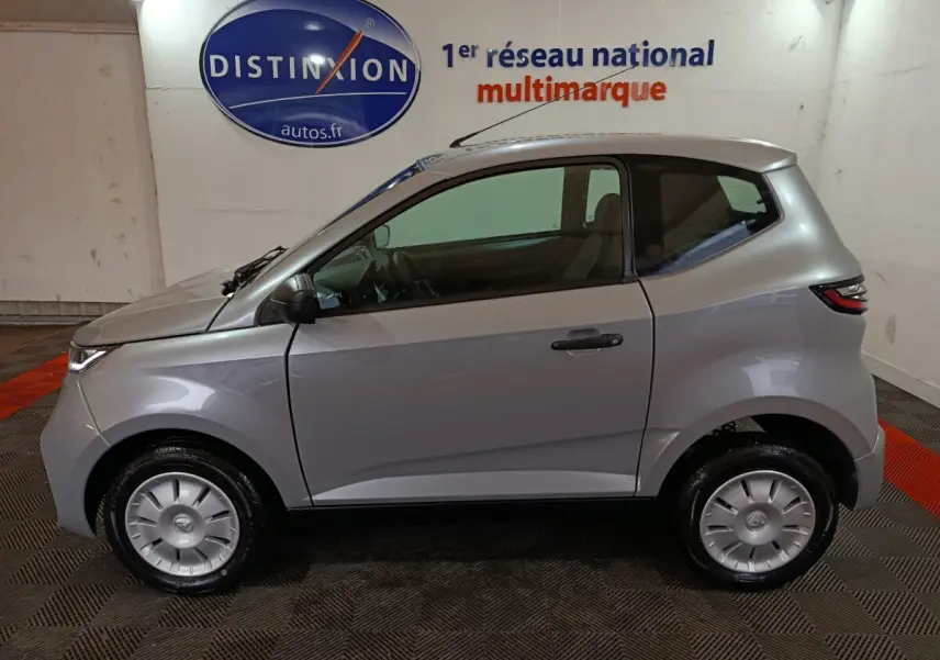 Profil côté gauche d'une Aixam Ambition 0.5 City Pack gris clair 2026 avec rétroviseur caméra conducteur en intérieur showroom.