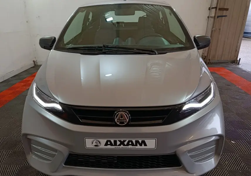 Vue avant d'une Aixam Ambition 0.5 City Pack gris clair avec phares allumés et rétroviseur caméra côté gauche.