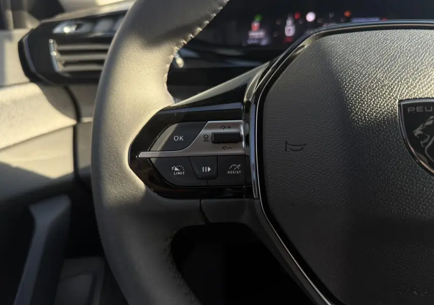 Gros plan sur le volant cuir noir de la Peugeot 308 2025, avec commandes intégrées côté gauche et logo central.