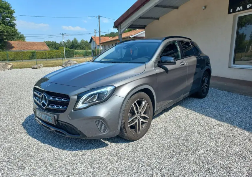 Mercedes GLA gris en 3/4 avant droit, avec calandre distinctive et jantes alliage, stationné sur gravier devant un bâtiment.