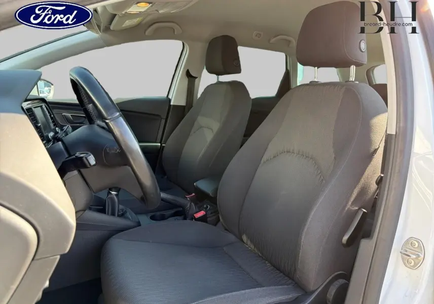 Vue intérieure côté conducteur de la SEAT LEON ST blanche, montrant les sièges tissu noirs et le volant multifonction.