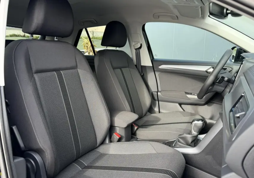 Vue intérieure côté gauche du Volkswagen T-Roc 1.5 TSI 2025, sièges tissu gris avec accoudoir central.