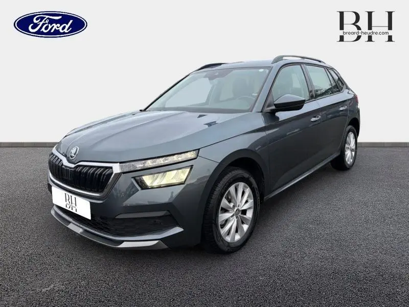 Vue 3/4 avant d'un Skoda Kamiq gris météore avec calandre chromée et phares LED allumés sur fond neutre.