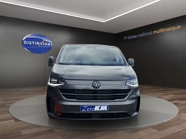 Vue avant d’un Volkswagen Transporter gris foncé 2026 avec phares LED et calandre noire dans un showroom.