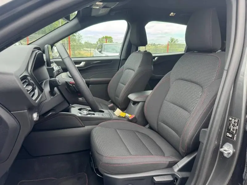 Vue intérieure côté conducteur du Ford Kuga 2023 gris Solar, sièges sport noirs avec surpiqûres rouges et volant cuir multifonction.