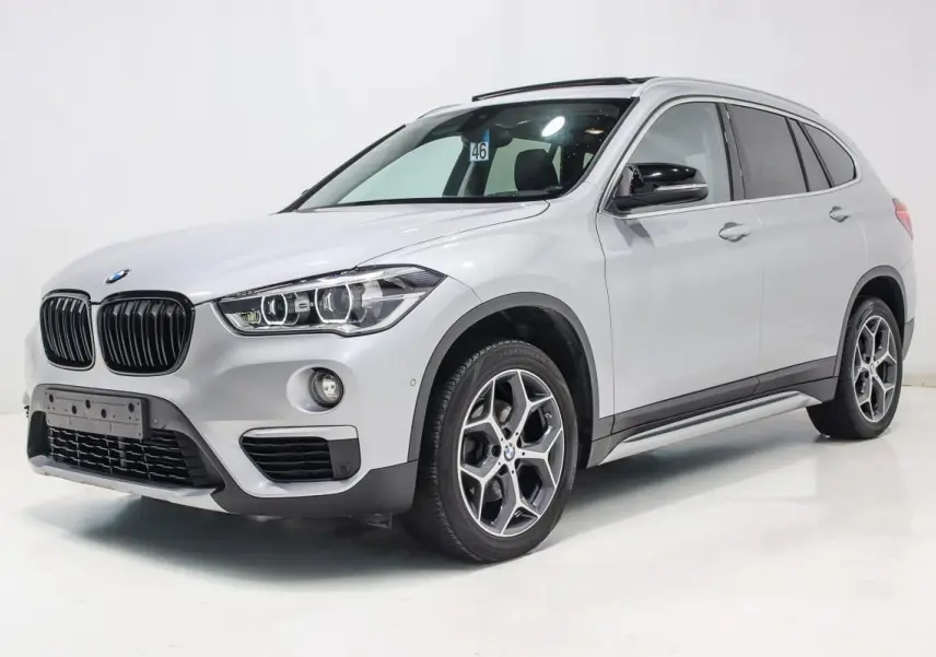 BMW X1 sDrive 18D argent métallisé en 3/4 avant droit avec toit ouvrant et attelage visible.