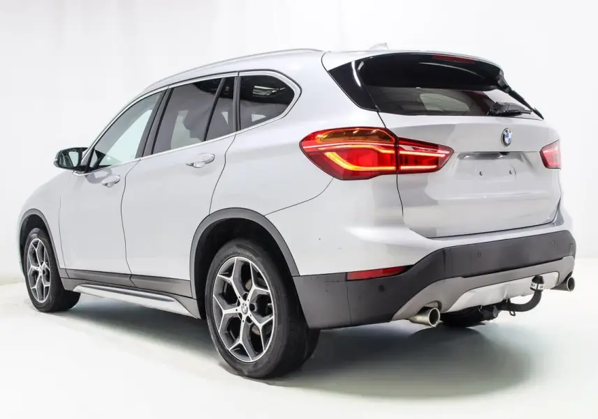 BMW X1 sDrive 18D argent métallisé vue 3/4 arrière droit avec attelage et feux arrière allumés.