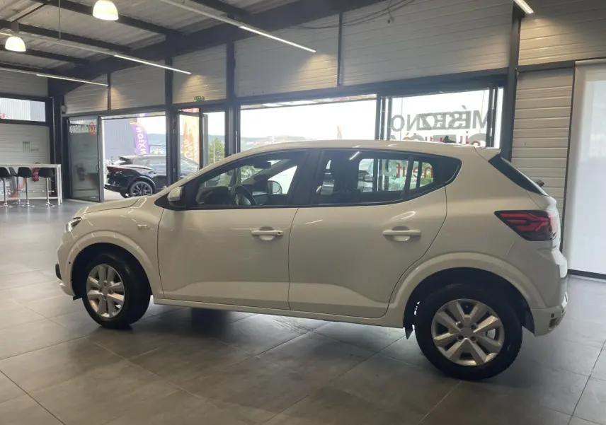 Profil côté gauche d'une Dacia Sandero blanche 2024 en showroom, avec jantes alliage et feux arrière LED distinctifs.