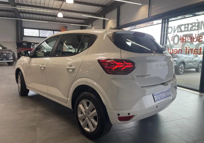 Vue 3/4 arrière droite d'une Dacia Sandero blanche 2024 en showroom, avec feux arrière LED et enjoliveurs 15 pouces.