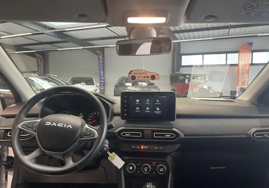 Vue intérieure avant du tableau de bord noir et volant DACIA d'une Sandero blanche avec écran tactile central allumé.