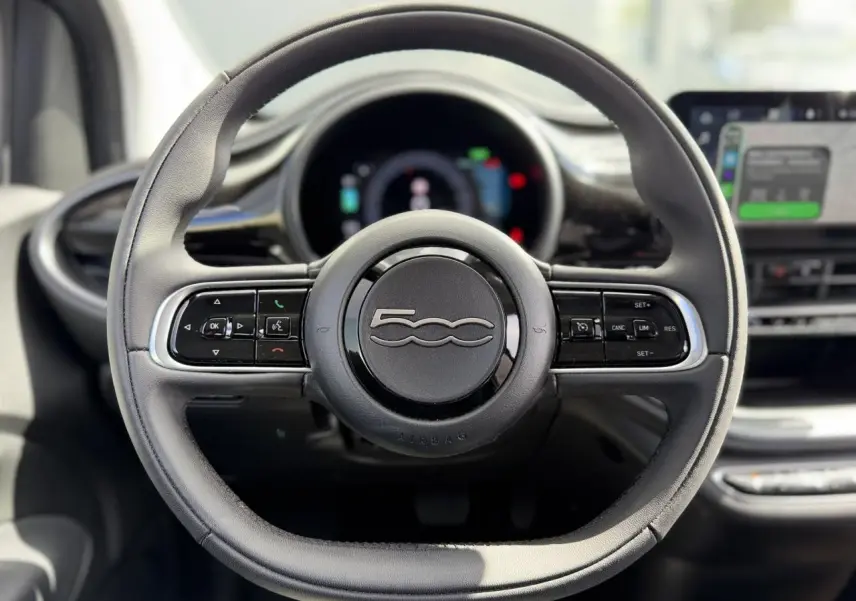 Volant noir Soft-Touch de la FIAT 500 électrique 2024 vu de face avec commandes intégrées et tableau de bord flou.