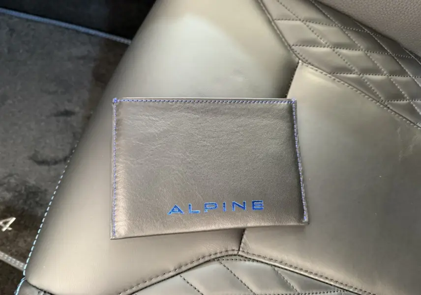 Gros plan sur un porte-documents en cuir noir avec logo Alpine bleu, posé sur un siège en cuir noir matelassé.