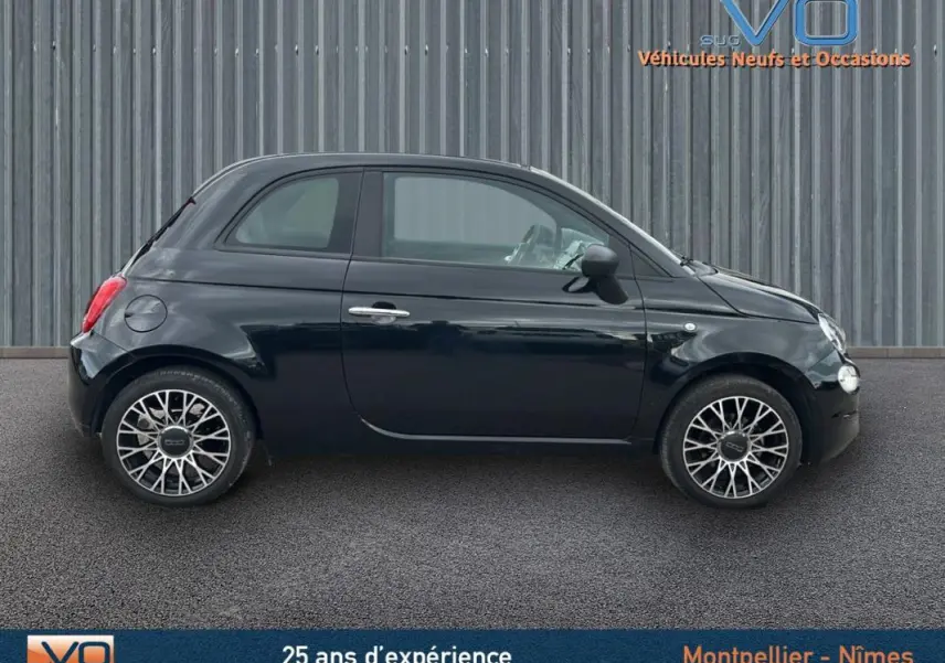 Profil côté gauche de la FIAT 500C noire 2024 avec roues alliage et roue de secours visible sur fond industriel.