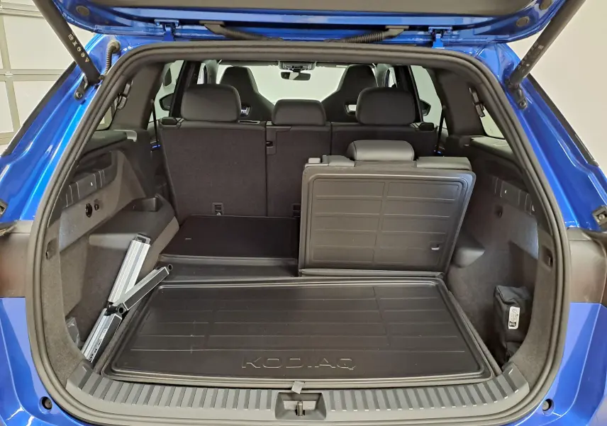 Coffre ouvert vu de l'arrière d'un Skoda Kodiaq Bleu RS 2025 avec banquette partiellement rabattue et tapis de coffre marqué KODIAQ.