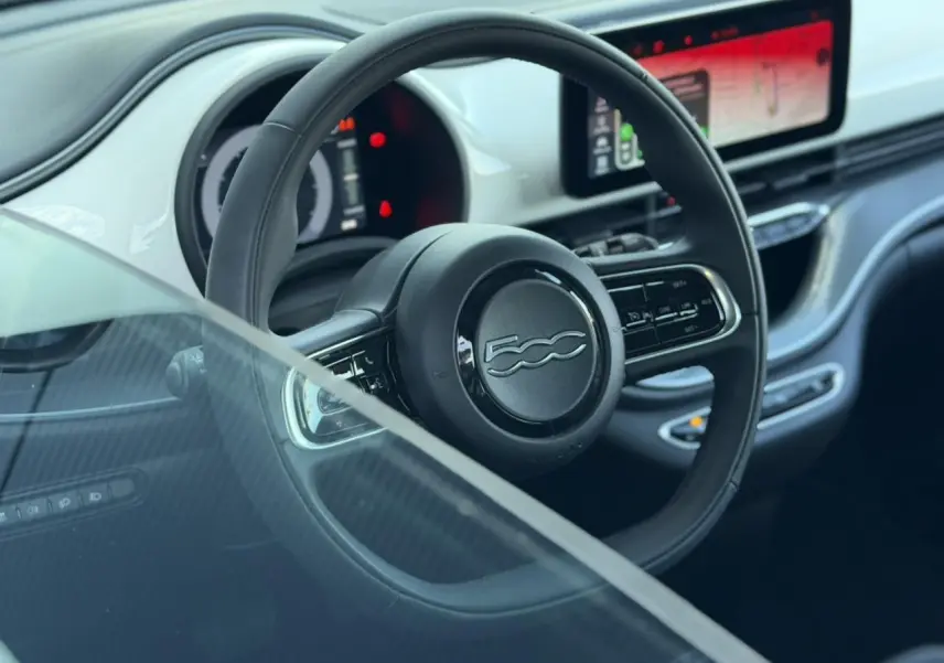 Vue intérieure centrée sur le volant noir et le tableau de bord numérique de la Fiat 500 électrique blanc Artic 2024.