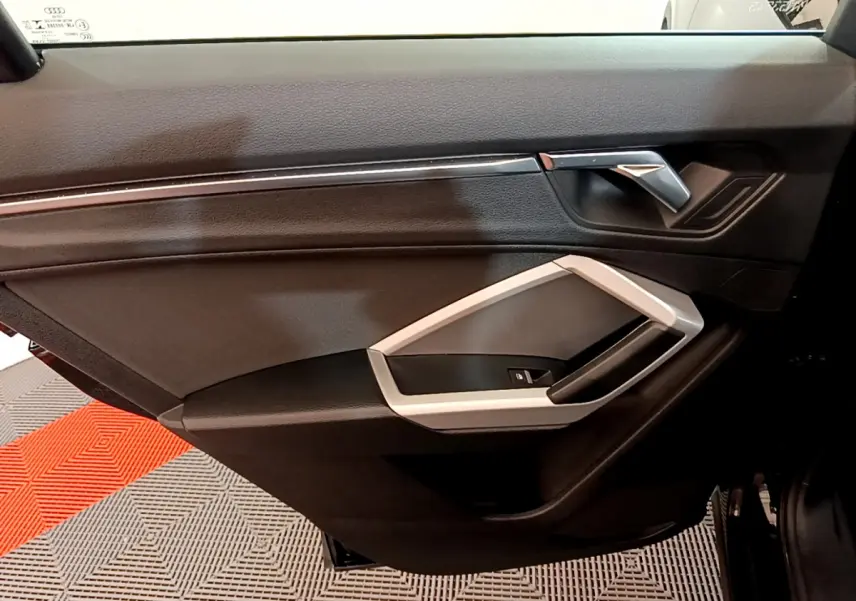 Vue intérieure côté gauche de la porte avant noire d’un Audi Q3 35 TDI 2021 avec poignée argentée et commandes fenêtres.