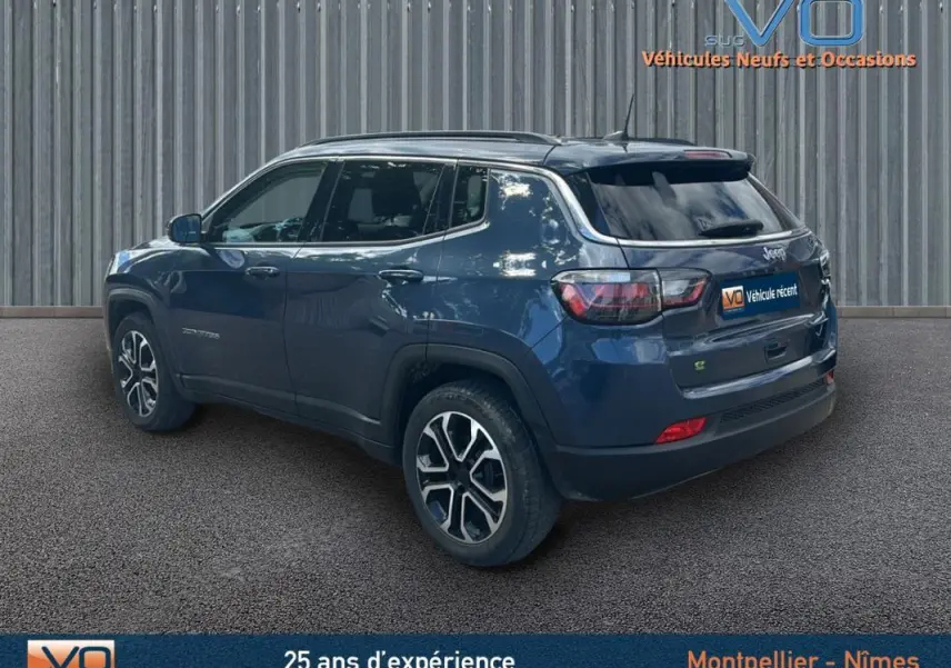 Vue 3/4 arrière droite d'un Jeep Compass bleu 2023 avec barres de toit et jantes 18 pouces, stationné devant un mur métallique.