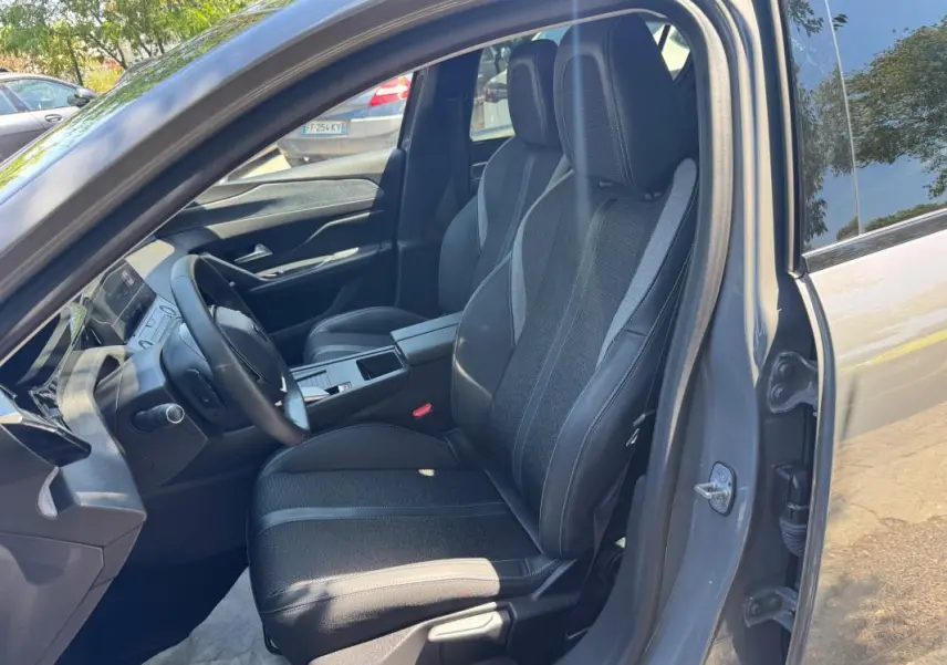 Vue intérieure côté conducteur d'une Peugeot 308 gris 2025, mettant en valeur les sièges noirs et l'accoudoir central.