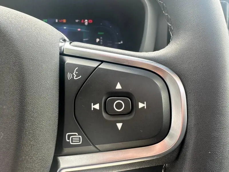Gros plan sur les commandes noires et argentées du volant du Volvo XC60 2024, avec affichage numérique flou en arrière-plan.