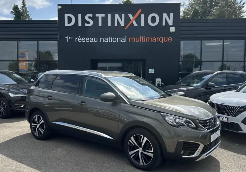 Peugeot 5008 gris amazonite en 3/4 avant droit, avec jantes alliage et rétroviseurs chromés, stationné devant un showroom.