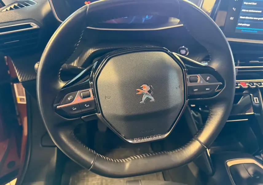 Vue rapprochée du volant compact en cuir noir avec logo Peugeot, tableau de bord et console centrale du 2008 orange 2020.