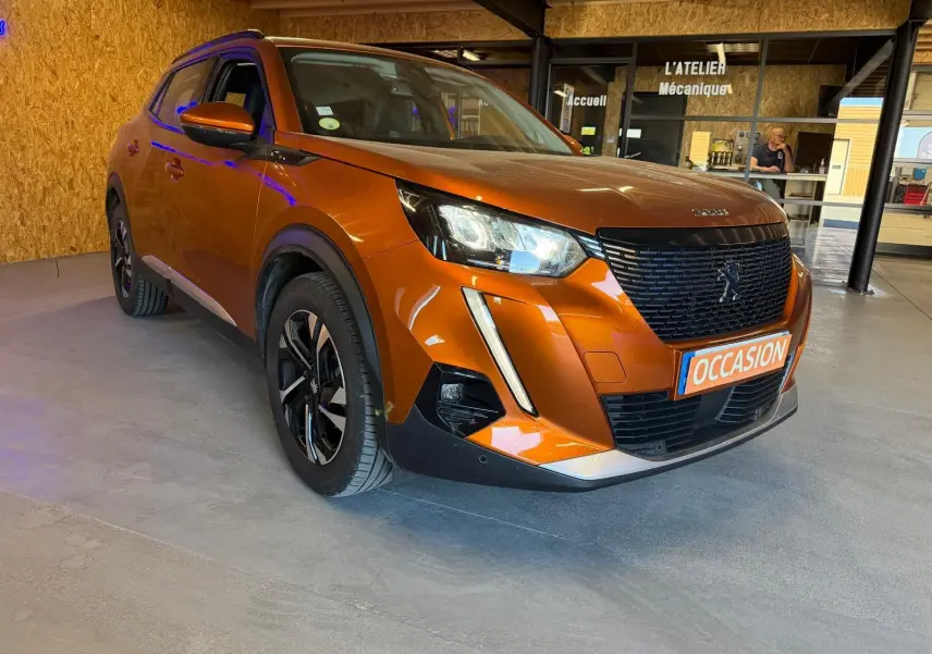 Peugeot 2008 orange vue 3/4 avant droit, avec calandre noire et jantes alliage bicolores, en intérieur showroom.