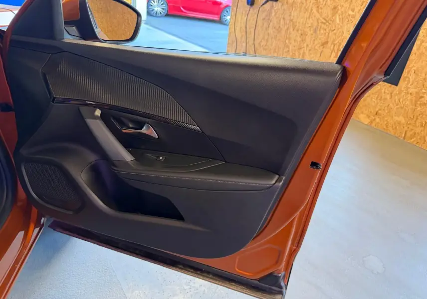 Vue intérieure de la porte avant gauche orange du Peugeot 2008 1.5 BlueHDi avec insert carbone et poignée chromée.