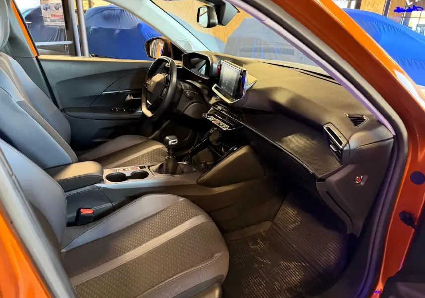 Vue intérieure côté conducteur du Peugeot 2008 orange, montrant la planche de bord et la sellerie tissu grise avec surpiqûres.