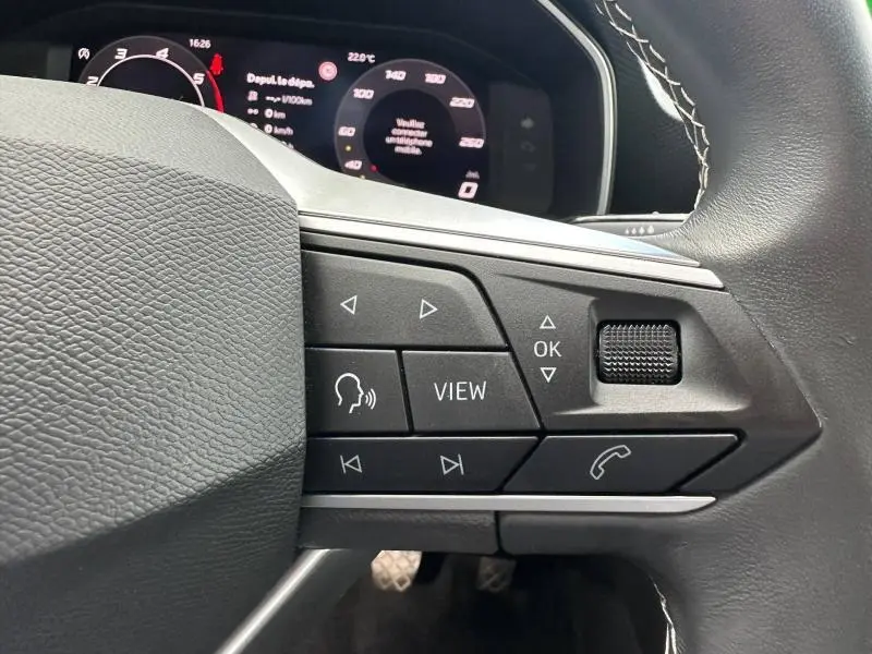 Gros plan sur les commandes multifonctions du volant cuir noir de la SEAT LEON 2025, avec tableau de bord numérique en arrière-plan.
