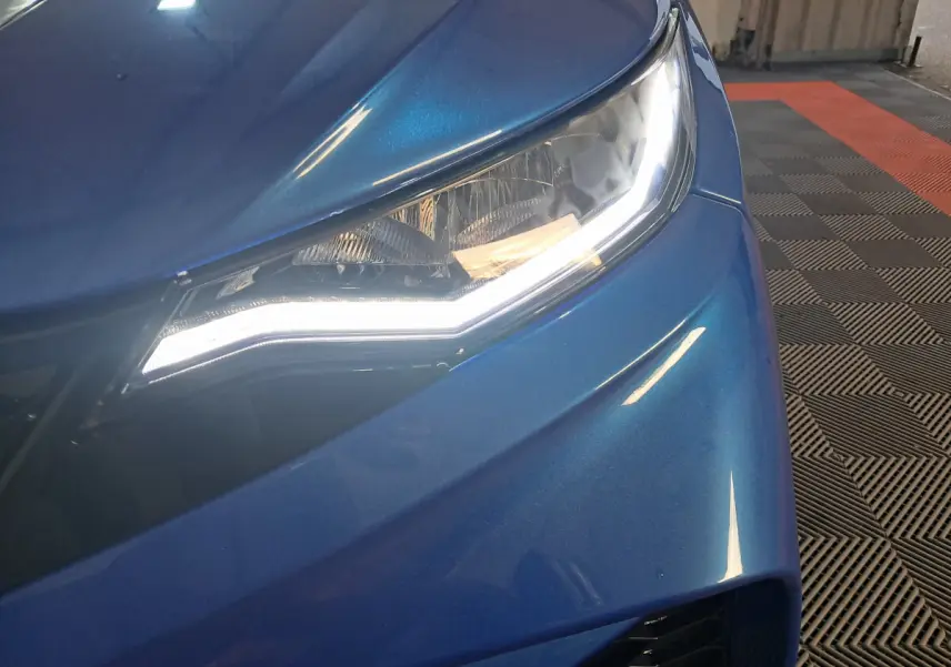 Gros plan sur le phare avant droit d'une Aixam Ambition 2025 bleu métallisé avec éclairage LED allumé.