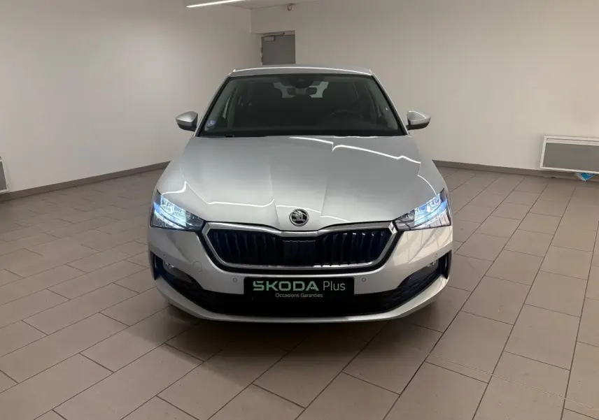 Vue frontale d'une Skoda Scala 1.0 TSI 110 DSG7 Business gris argent avec phares allumés en intérieur showroom.