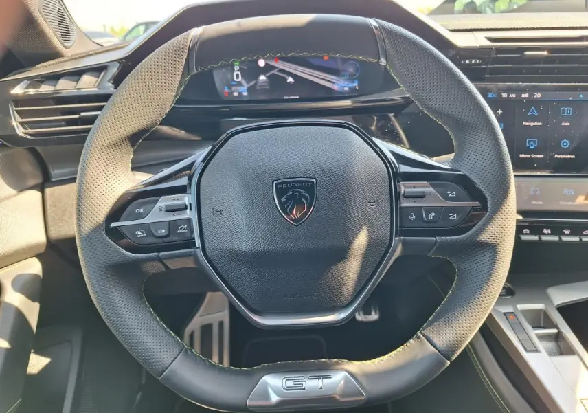 Vue rapprochée du volant cuir noir avec logo Peugeot et badge GT, tableau de bord numérique et écran tactile en arrière-plan.