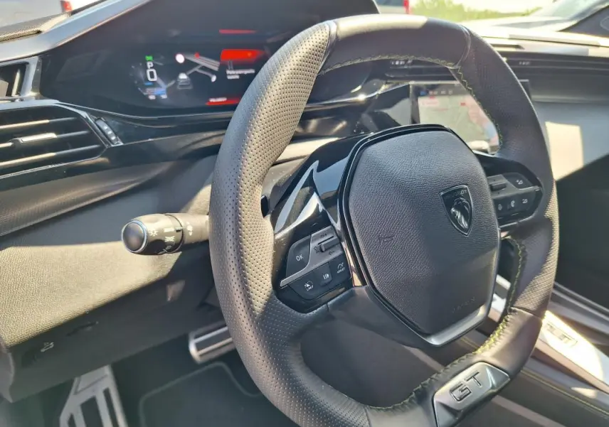 Vue intérieure rapprochée du volant GT en cuir perforé et du cockpit numérique de la Peugeot 308 SW Hybrid 145 noir.