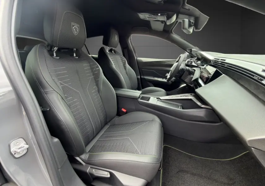 Vue intérieure côté conducteur de la Peugeot 308 GT 2026 gris clair, sièges noirs avec logo lion brodé et tableau de bord moderne.