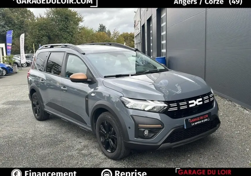 Vue 3/4 avant d’un Dacia Jogger Extreme+ Hybrid 140 gris avec rétroviseurs bronze et jantes noires, stationné devant un bâtiment.