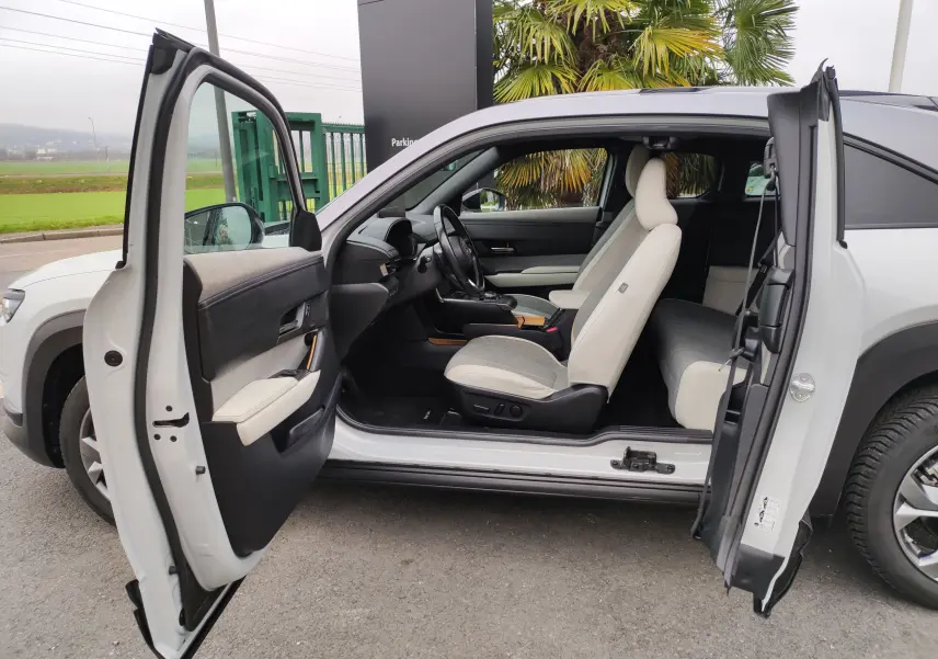 Vue latérale gauche ouverte d'une Mazda MX-30 gris clair 2023, mettant en valeur l'intérieur clair et moderne avec sièges bicolores.